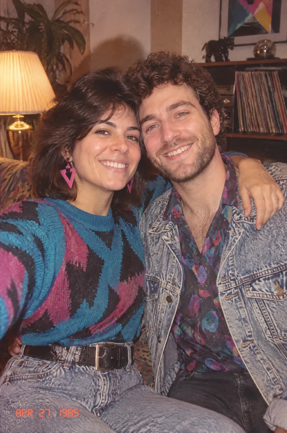 80s flashback snapshot of a couple posing close together in a living room with record shelves, flash glare and an orange date stamp