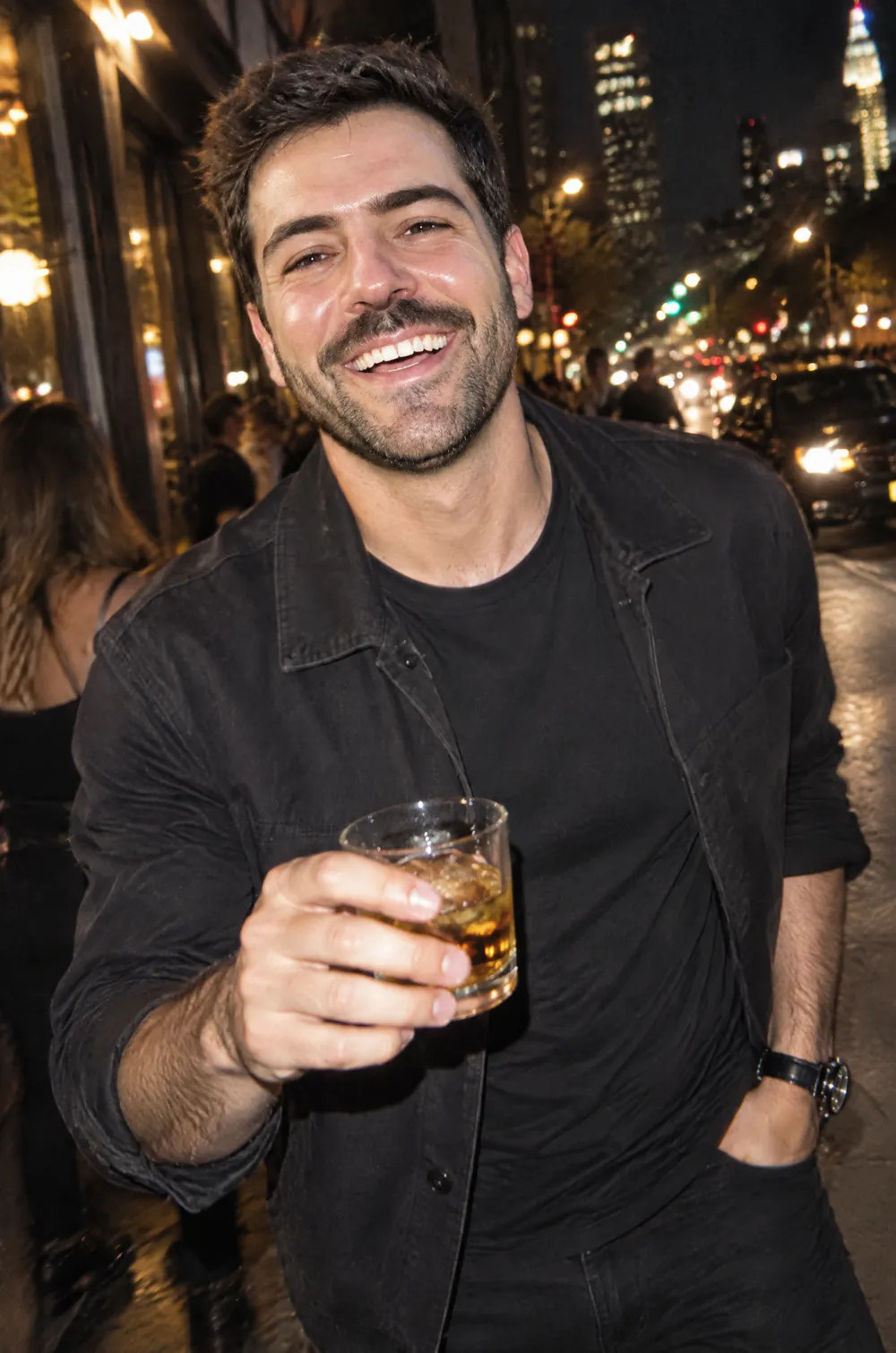 Night flash photo of a bearded man in a dark jacket raising a glass on a city street, with glossy flash highlights