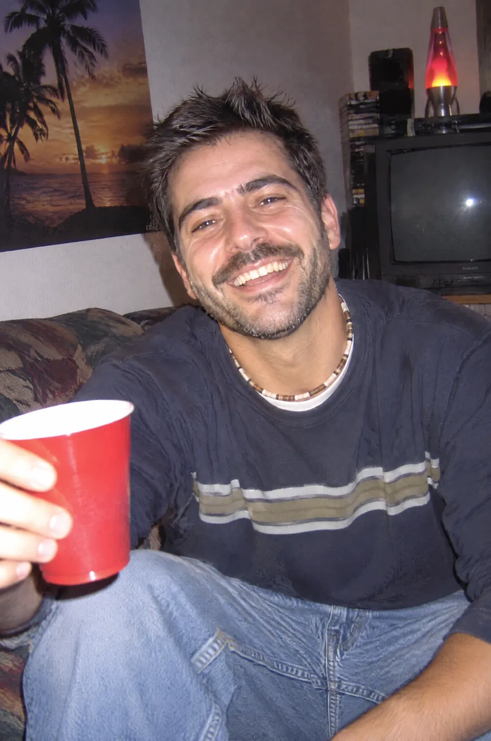 Y2K digital camera snapshot of a smiling man in a striped sweater holding a red cup, with flash glare and digital noise